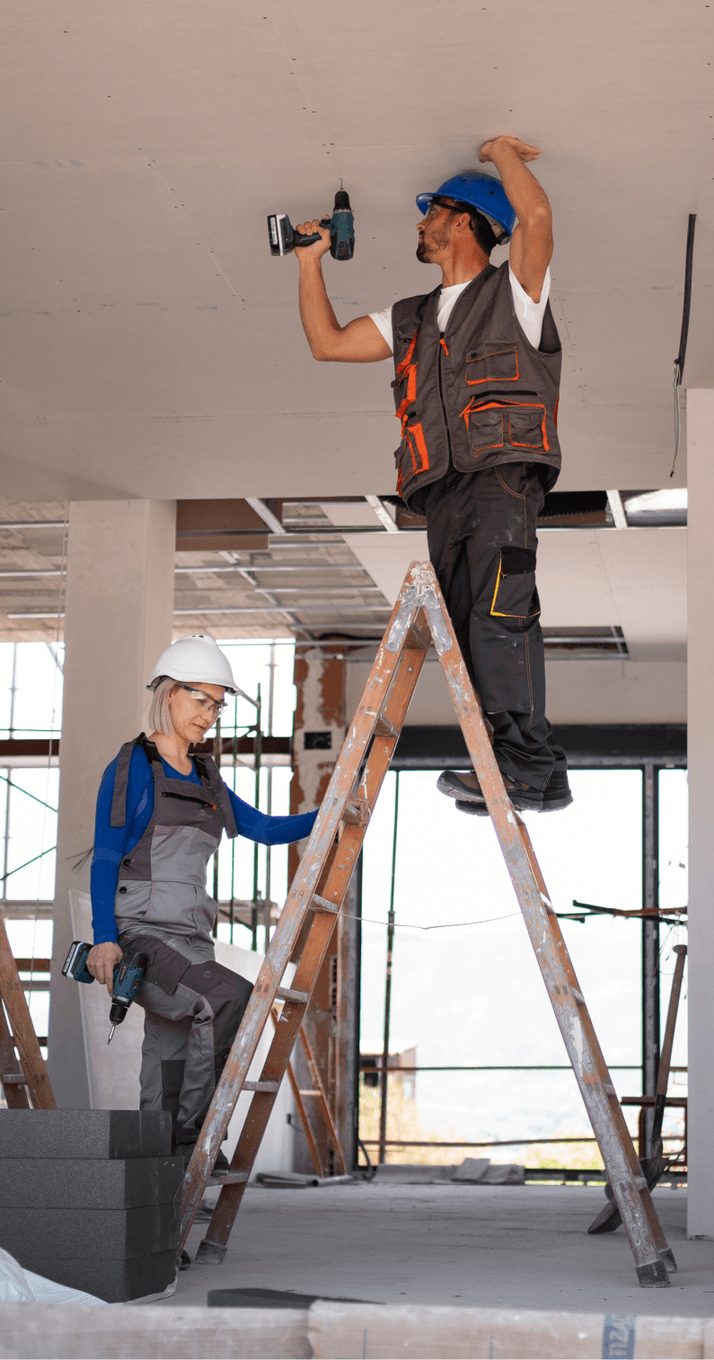 construction-img A male worker on a ladder drills into a ceiling, while a female supervisor observes in a construction site. both wear hard hats and safety vests.