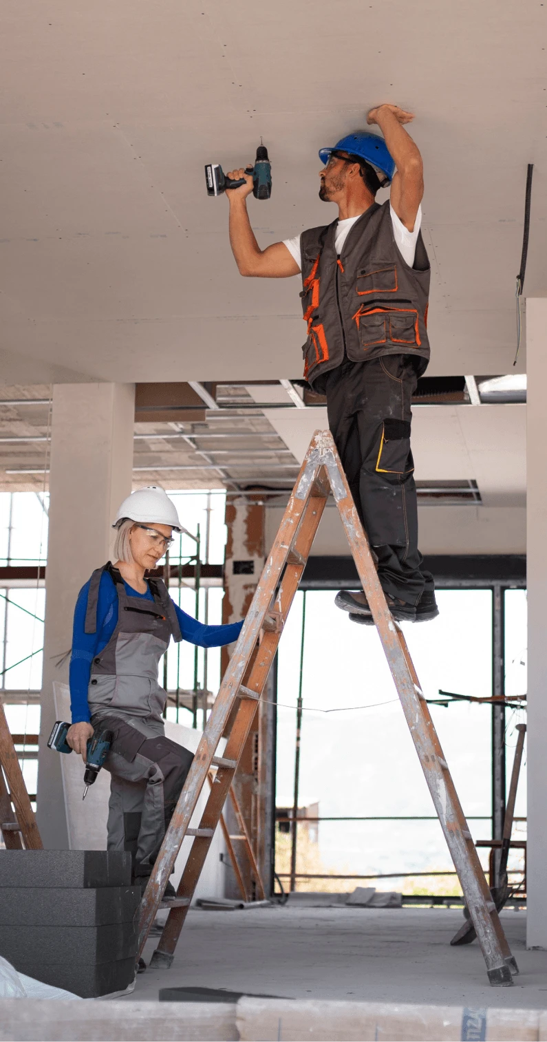 A male worker on a ladder drills into a ceiling, while a female supervisor observes in a construction site. both wear hard hats and safety vests.