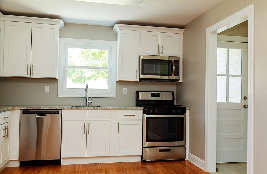 A modern kitchen with white cabinetry, stainless steel appliances, and a granite countertop. there's a window above the sink and a doorway on the right.