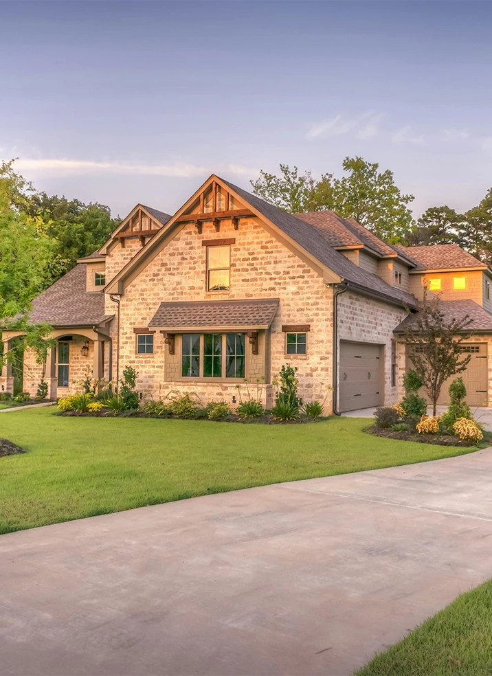 Two-story brick and stone house with a gabled roof, manicured lawn, and a concrete driveway, under a clear sky at dusk.